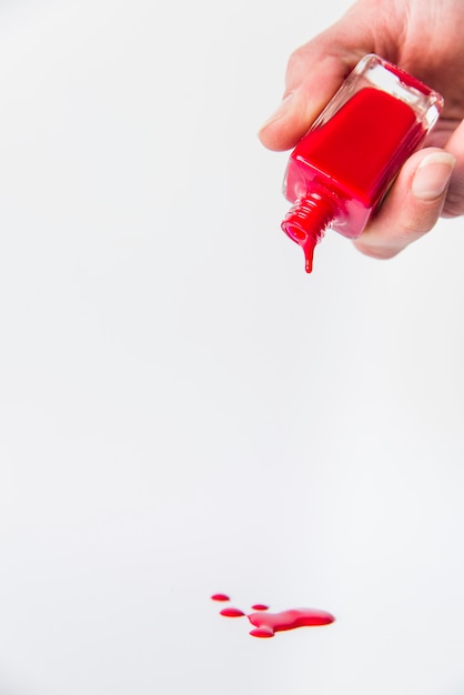 Primo piano della mano che versa la bottiglia di smalto per unghie rossa sul contesto bianco