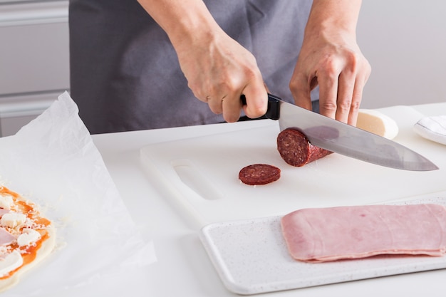 Primo piano della mano della donna che taglia le fette del salame con il coltello tagliente sul tagliere