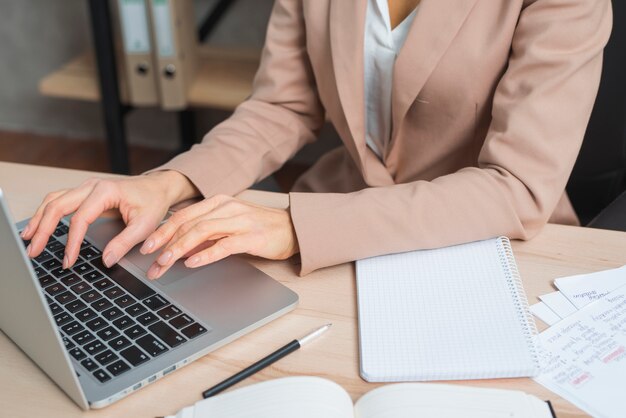 Primo piano della mano della donna di affari che scrive sul computer portatile con la penna; diario e blocco note a spirale sul tavolo di legno