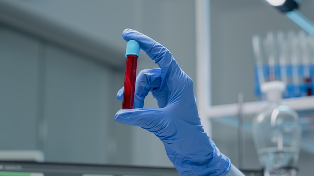 Primo piano della mano specialistica con guanti che tengono vacutainer con soluzione rossa o sangue nel laboratorio di ricerca. Strumento di scienza del vetro con liquido in bicchiere trasparente utilizzato per l'innovazione