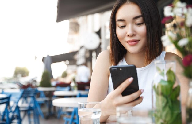 Primo piano della moderna donna asiatica seduta al caffè sul marciapiede con il telefono cellulare Ragazza che legge un messaggio di testo sullo smartphone mentre ci si rilassa in città