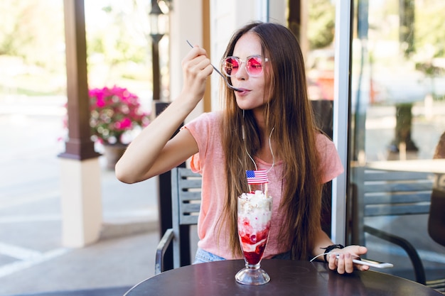 Primo piano della ragazza carina seduta in un caffè a mangiare il gelato con la ciliegina sulla torta con un cucchiaio. Indossa pantaloncini di jeans, top rosa e sorrisi. Ha gli occhiali rosa. Ascolta la musica sullo smartphone