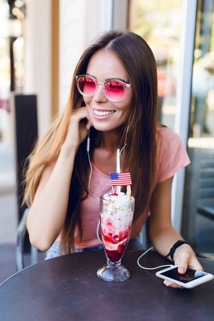 Primo piano della ragazza carina seduta in un caffè a mangiare il gelato con la ciliegina sulla torta. Indossa un top rosa e occhiali rosa. Ascolta la musica sullo smartphone e sorride. Ha lunghi capelli scuri