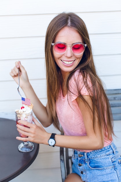 Primo piano della ragazza carina seduta su una sedia a mangiare il gelato con la ciliegina sulla torta con un cucchiaio. Indossa pantaloncini di jeans, top rosa e sorrisi. Ha gli occhiali rosa