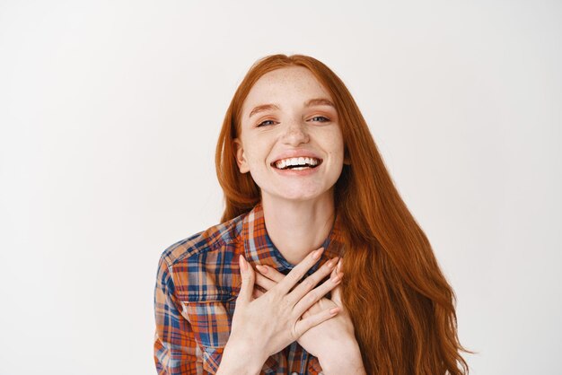 Primo piano della ragazza felice dello zenzero con la pelle pallida e gli occhi azzurri che sorridono grati esprimendo gratitudine e gioia che si tengono per mano sul cuore che stanno sopra fondo bianco