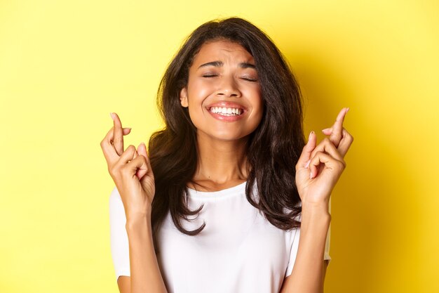 Primo piano della ragazza nervosa afro-americana, incrociando le dita per buona fortuna e in attesa di risultati importanti, esprimere un desiderio, in piedi su sfondo giallo.