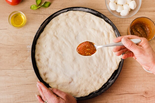 Primo piano della salsa della holding della mano di una donna in cucchiaio per l&#39;applicazione sulla pasta della pizza