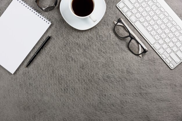 Primo piano della tazza di caffè bianco; tastiera; orologio da polso; penna; blocco note a spirale; occhiali da vista e tastiera sulla scrivania