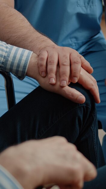 Primo piano delle mani dell'infermiera e del paziente anziano durante la visita medica in casa di cura. Assistente medico che dà supporto e conforta il vecchio in sedia a rotelle. Specialista che aiuta la persona disabile