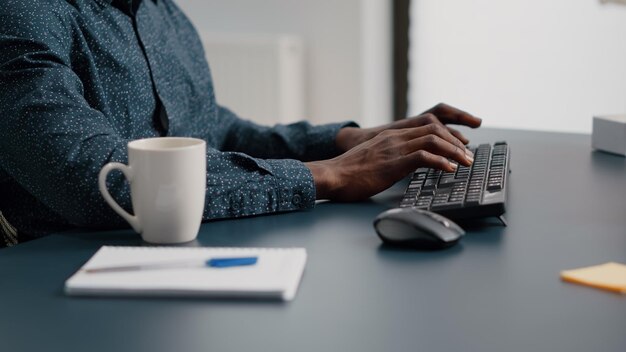 Primo piano delle mani dell'uomo afroamericano che digitano sulla tastiera del computer in soggiorno, utilizzando la comunicazione web online internet dall'ufficio di casa. Lavoratore remoto che lavora da casa