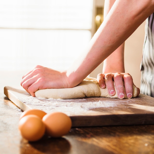 Primo piano delle mani della donna che impasta la pasta sul tagliere con tre uova sulla tavola di legno