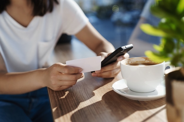 Primo piano delle mani delle donne che si appoggia sul tavolino da caffè, che tiene il telefono cellulare e la carta di credito di plastica.