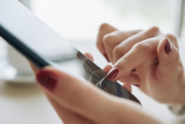 Primo piano delle mani femminili con il manicure rosso scuro dell'unghia che per mezzo dello smartphone