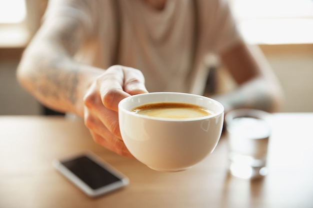 Primo piano delle mani maschili che propongono una tazza di caffè, seduti al tavolo con lo smartphone. Navigare, fare acquisti online, lavorare. Istruzione, freelance, arte e concetto di business. Potabile. Bevanda aromatica calda.