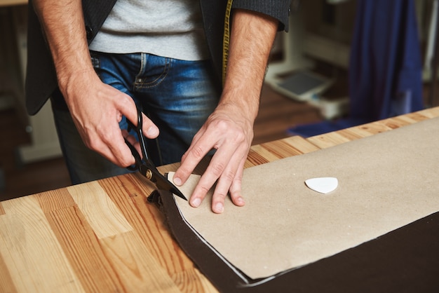 Primo piano dettagliato di giovane stilista maschio di successo che lavora alla nuova collezione di abiti, ritagliando le parti del vestito dal tessuto, creando passo dopo passo il capolavoro.