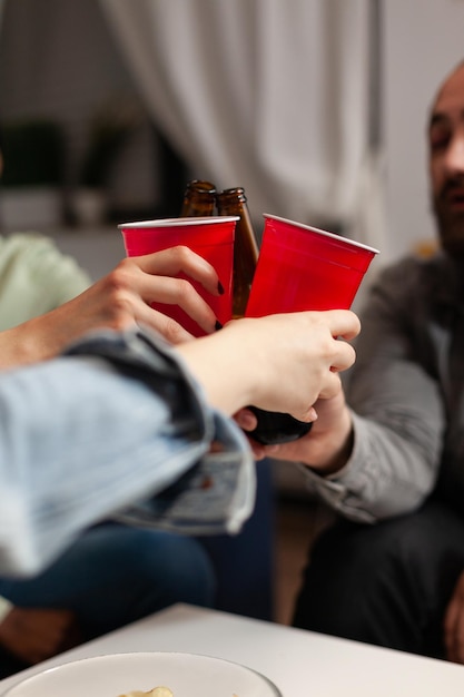 Primo piano di amici che tintinnano bicchiere di plastica con la birra durante la festa di fine settimana godendo di trascorrere del tempo insieme. Gruppo multietnico di persone che bevono alcolici in soggiorno. Concetto di amicizia