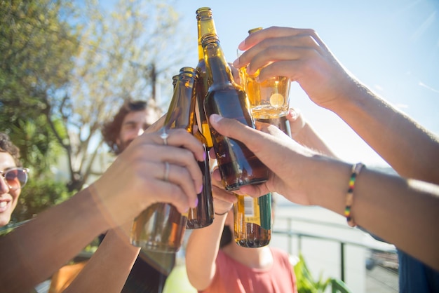 Primo piano di amici contenuti che tintinnano bottiglie di birra e bicchieri. Persone di diverse nazionalità che allevano bottiglie, ridono, festeggiano il compleanno. Festa, concetto di amicizia