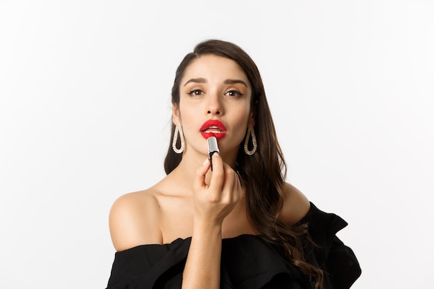 Primo piano di bella donna che applica rossetto rosso sulle labbra, che guarda l'obbiettivo come specchio, in piedi in abito nero su sfondo bianco.