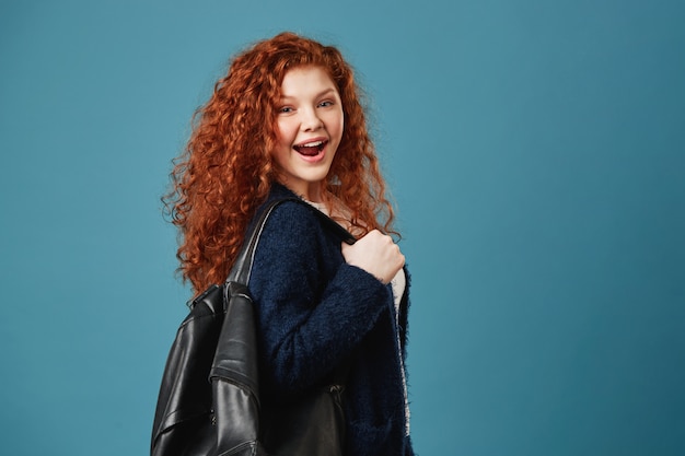 Primo piano di bella donna felice studente zenzero con capelli ondulati tenendo lo zaino con la mano, sorridendo felicemente.