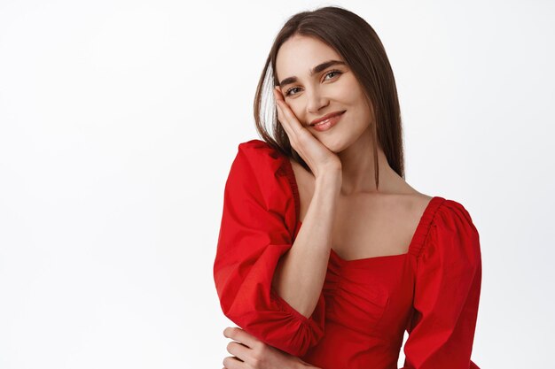 Primo piano di bella giovane donna in elegante abito rosso, abito romantico glamour per un appuntamento o una festa serale, guardando in modo civettuolo e sorridendo alla telecamera, sfondo bianco