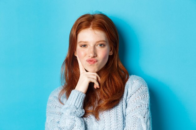 Primo piano di bella ragazza rossa che pensa, labbra pucker e fissando la fotocamera t pensierosa, facendo una scelta, in piedi su sfondo blu.