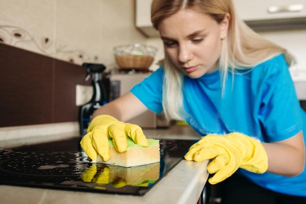 Primo piano di donna che indossa un guanto protettivo con straccio per la pulizia della stufa elettrica a casa cucina Bella ragazza che lava la superficie nera lucida della mano superiore della cucina con schiuma sulla spugna Concetto di lavoro domestico