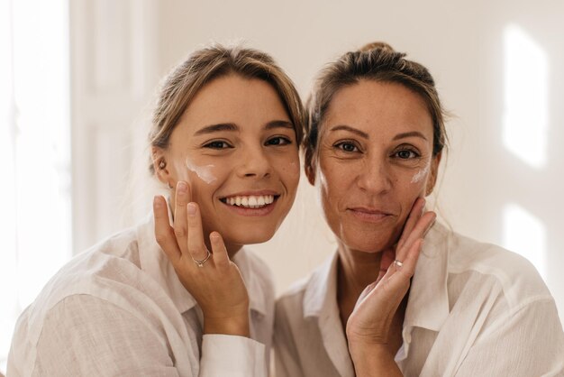 Primo piano di due donne caucasiche carine che si spalmano il viso con crema e guardano la fotocamera su sfondo bianco. Concetto di bellezza e giovinezza