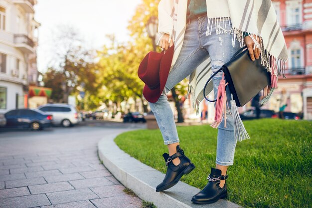 Primo piano di gambe di donna che indossa stivali di pelle nera, jeans, tendenze primaverili di calzature, borsa della holding