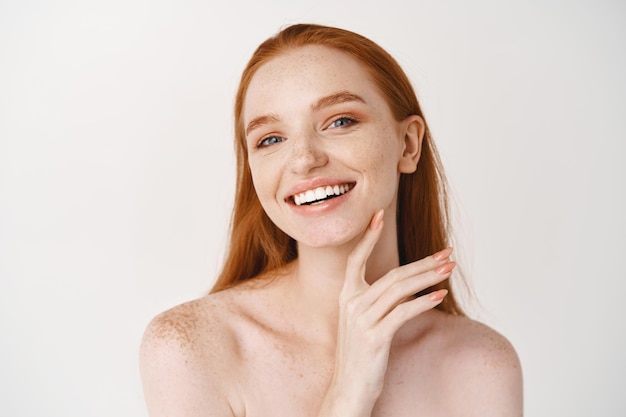 Primo piano di giovane bella donna rossa che sorride davanti, tocca la pelle pulita perfetta sul viso e sembra felice, in piedi nuda sul muro bianco