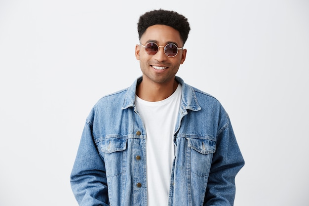 Primo piano di giovane bello allegro alla moda uomo dalla pelle scura con acconciatura afro in camicia bianca sotto giacca di jeans e occhiali da sole sorridente con i denti, guardando a porte chiuse con felice expressio