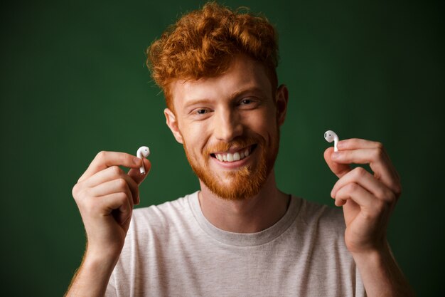 Primo piano di giovane sorridente sorridente riccio barbuto giovane in maglietta bianca, mostrando airpods