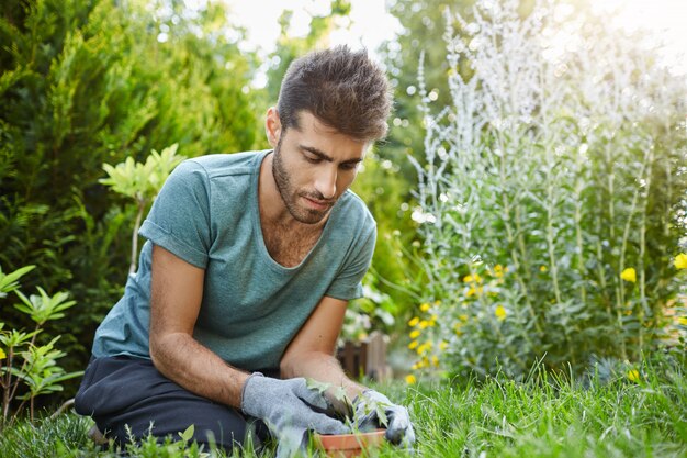 Primo piano di giovane uomo barbuto serio in maglietta blu e guanti concentrati a lavorare in giardino, piantando germogli in vaso di fiori. Giardiniere che trascorre la giornata nella sua casa di campagna