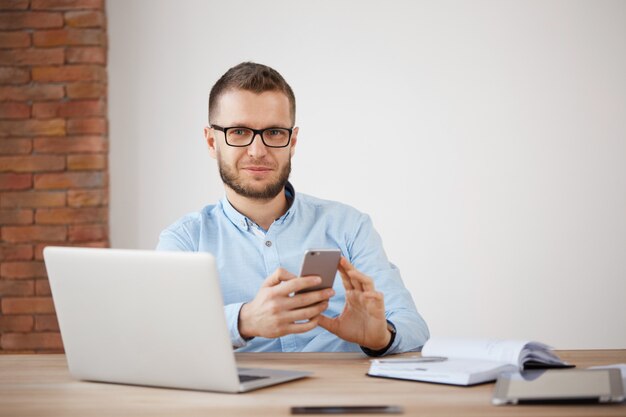 Primo piano di maturo maschio con la barba lunga manager in bicchieri e camicia seduto in ufficio, lavorando su un computer portatile