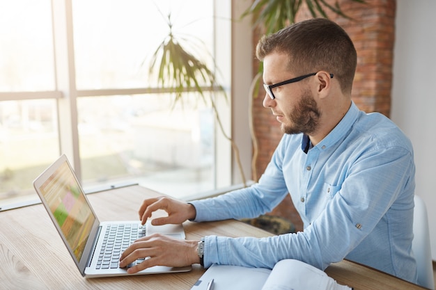 Primo piano di maturo serio barbuto maschio caucasico direttore della società seduto in un grande ufficio confortevole, guardando attraverso l'elenco dei clienti sul computer portatile, prendendo nota in taccuino, spendendo morni produttivi