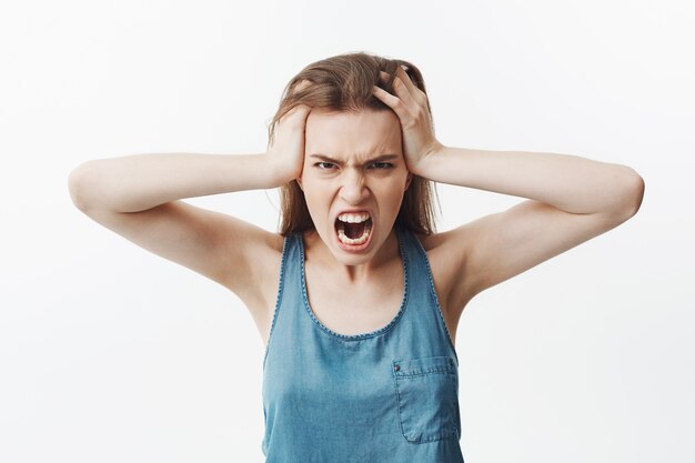 Primo piano di negativo giovane ragazza studentessa europea con lunghi capelli scuri in testa camicia blu con le mani, con espressione faccia arrabbiata e arrabbiata, urlando a gran voce