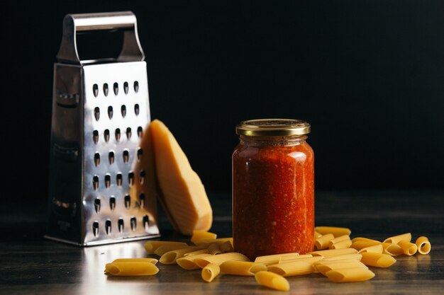 Primo piano di pasta di penne e un barattolo di salsa sul tavolo con formaggio e una grattugia sullo sfondo