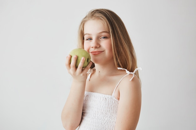 Primo piano di ragazza divertente con lunghi capelli biondi tenendo la mela nelle mani con espressione soddisfatta, andando a pranzo sano hava a scuola.