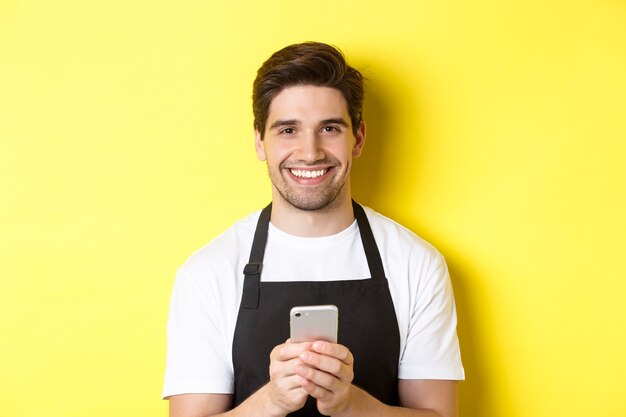 Primo piano di un bel barista che invia un messaggio sul cellulare, sorride felice, in piedi su sfondo giallo