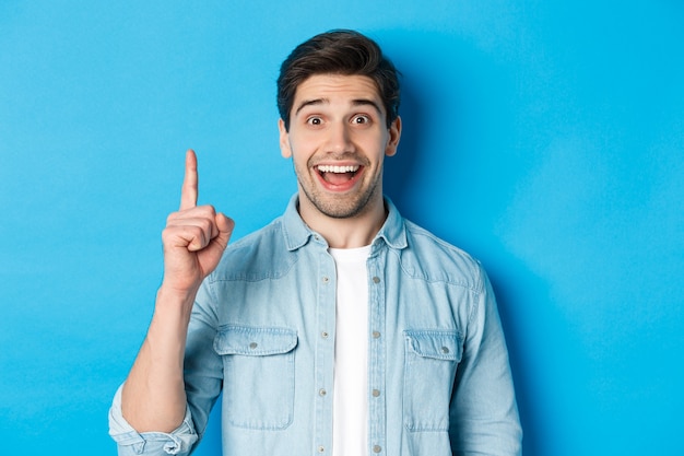 Primo piano di un bel ragazzo barbuto che sorride, mostrando il dito numero uno, in piedi su sfondo blu
