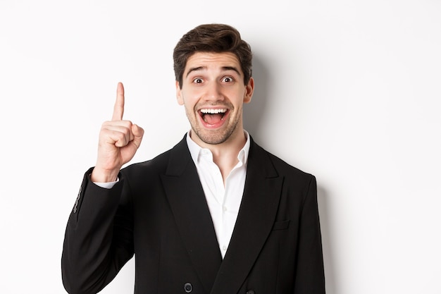 Primo piano di un bell'uomo d'affari in abito nero, sorridente stupito, che mostra il numero uno, in piedi su sfondo bianco
