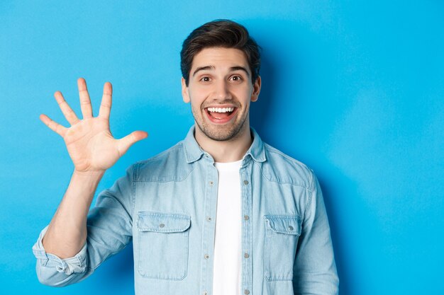 Primo piano di un bell'uomo sorridente, che mostra le dita numero cinque, in piedi su sfondo blu.