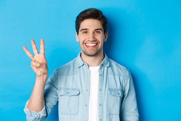 Primo piano di un bell'uomo sorridente, che mostra le dita numero tre, in piedi su sfondo blu