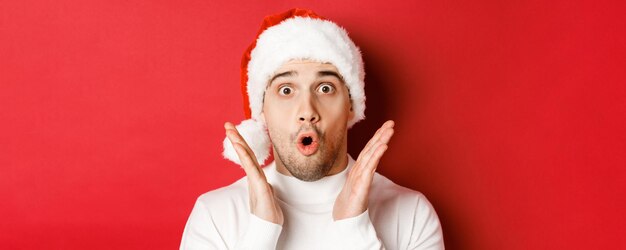 Primo piano di un bell'uomo stupito con cappello da Babbo Natale e maglione bianco, alzando le mani e guardando impressionato, in piedi su sfondo rosso