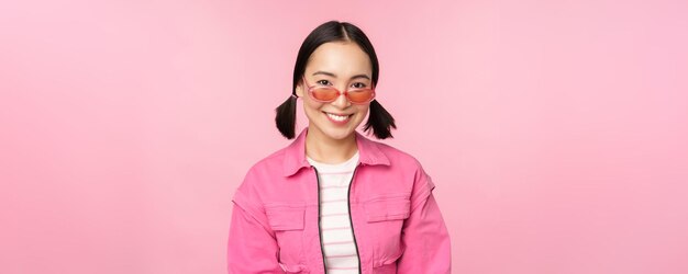 Primo piano di un bellissimo modello femminile asiatico in occhiali da sole alla moda in posa su sfondo rosa in uno spazio di copia vestito alla moda