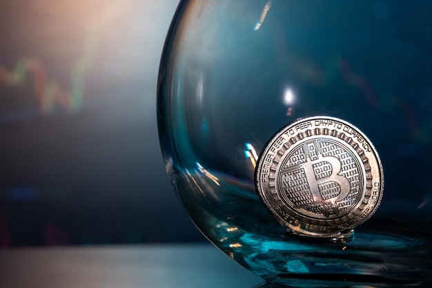Primo piano di un Bitcoin d'argento su una superficie riflettente blu in un bicchiere e l'istogramma della valuta