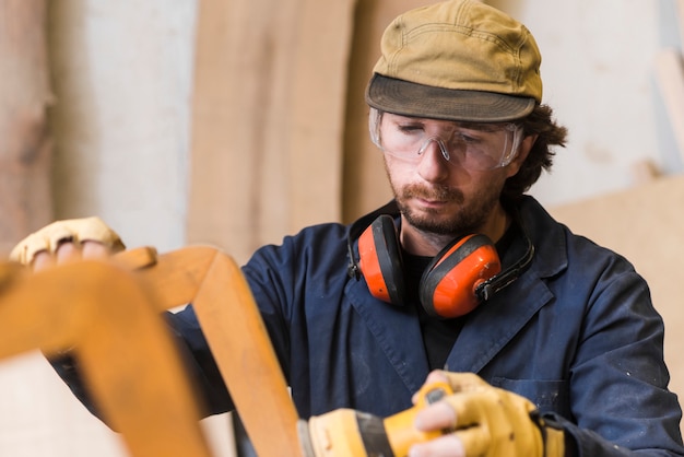 Primo piano di un carpentiere maschio che indossa occhiali di protezione e protezione per le orecchie intorno al collo usando la levigatrice elettrica