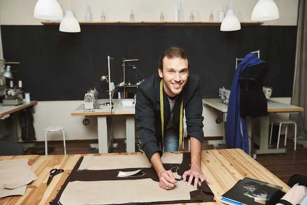 Primo piano di un giovane allegro attraente maschio designer di abbigliamento con acconciatura alla moda in tuta lavorando sulla nuova collezione nel suo laboratorio, tagliando le parti di vestiti.