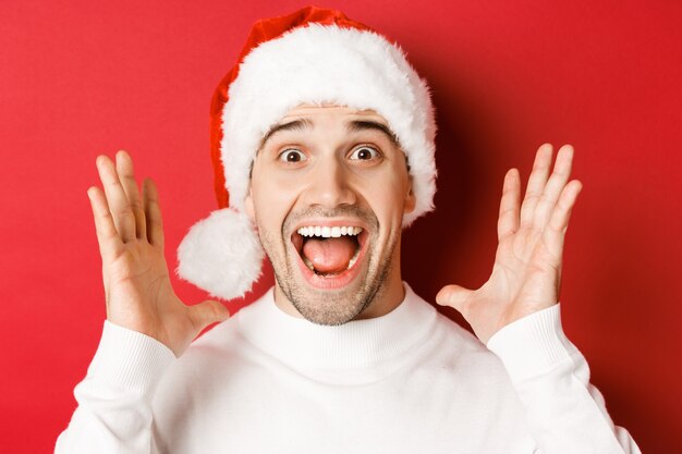 Primo piano di un giovane felice con un cappello da Babbo Natale, che fa un grande annuncio di Natale, sorride stupito, in piedi su uno sfondo rosso