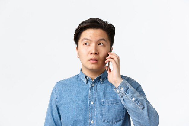 Primo piano di un ragazzo asiatico indeciso che si distanzia durante la telefonata, ha una conversazione e distoglie lo sguardo confuso, in piedi su uno sfondo bianco. Uomo che tiene lo smartphone vicino all'orecchio, ordina la consegna del cibo.