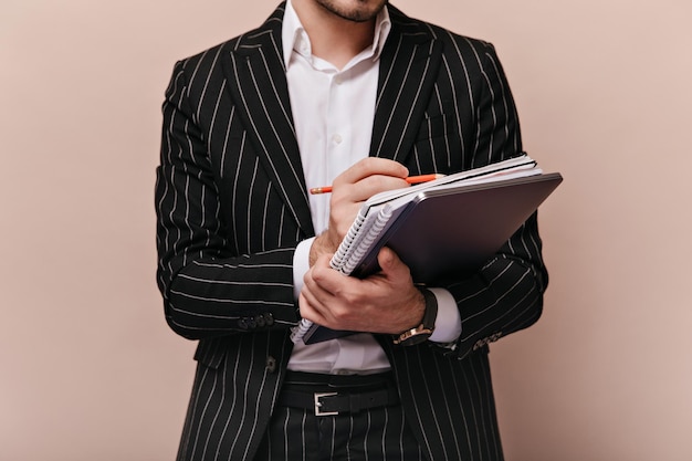 Primo piano di un uomo alla moda in camicia bianca e abito a righe nere che tiene cartelle e appunti con la penna in posa isolate su sfondo beige